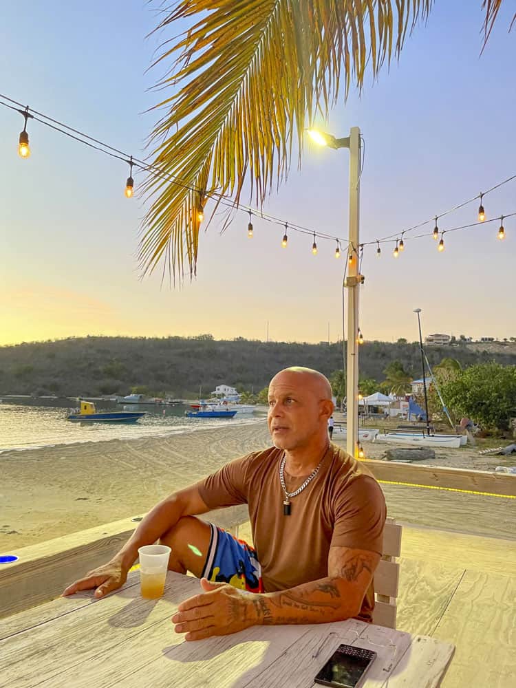 A man sitting at a table on the beach with a drink.